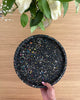 Black speckled tray held by a hand on a wooden surface with flowers on the side.